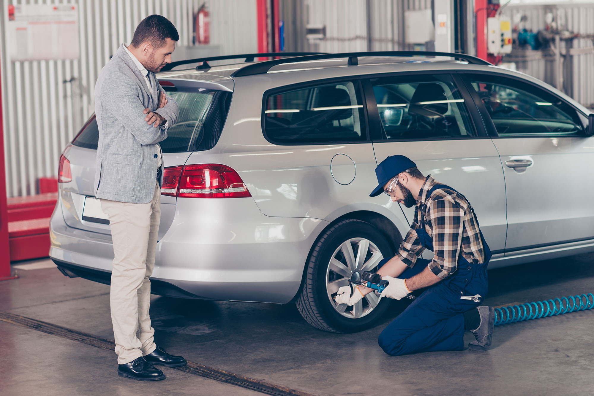 Bearded specialist technician expert in protective glasses and blue uniform is analyzing tire pressure of silver car on hardware, businessman in classy suit is standing near with crossed hands